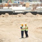 Dois trabalhadores da construção usando equipamentos de segurança conversam e analisam planos em um canteiro de obras.