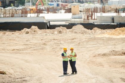 Dois trabalhadores da construção usando equipamentos de segurança conversam e analisam planos em um canteiro de obras.