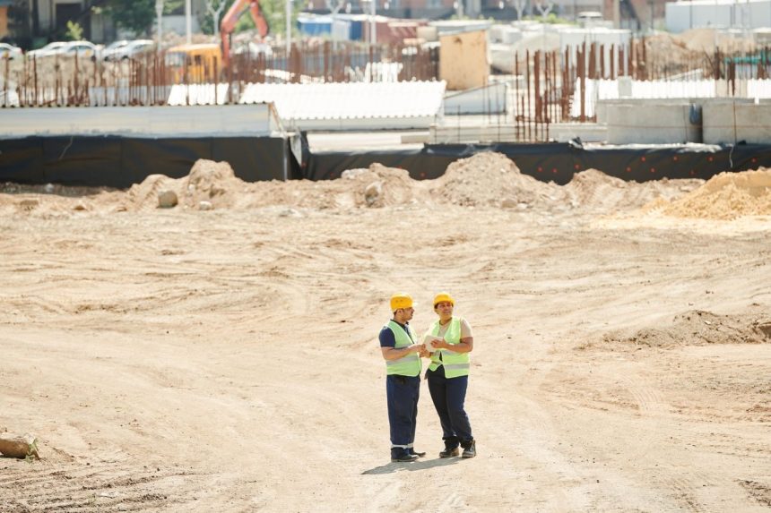 Dois trabalhadores da construção usando equipamentos de segurança conversam e analisam planos em um canteiro de obras.
