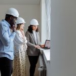 Três engenheiros usando capacetes analisam plantas de construção em um laptop em um ambiente interno.