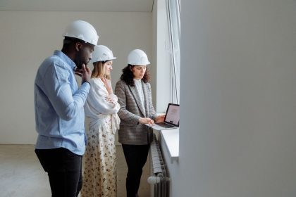 Três engenheiros usando capacetes analisam plantas de construção em um laptop em um ambiente interno.