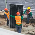Trabalhadores usando equipamentos de proteção individual instalam painéis solares em um canteiro de obras.